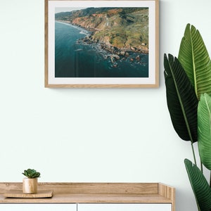 May include: A framed photograph of a rugged coastline with green hills and a blue ocean. The photo is in a light wood frame and is hanging on a white wall.