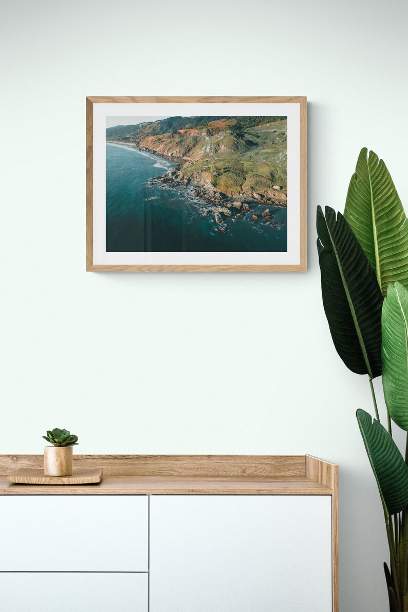May include: A framed photograph of a rugged coastline with green hills and a blue ocean. The photo is in a light wood frame and is hanging on a white wall.