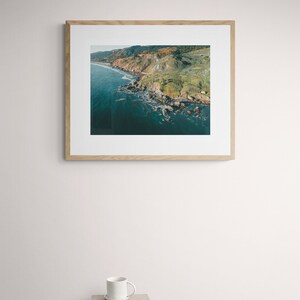 May include: A framed photograph of a rocky coastline with green vegetation and blue water. The photo is in a light wood frame.