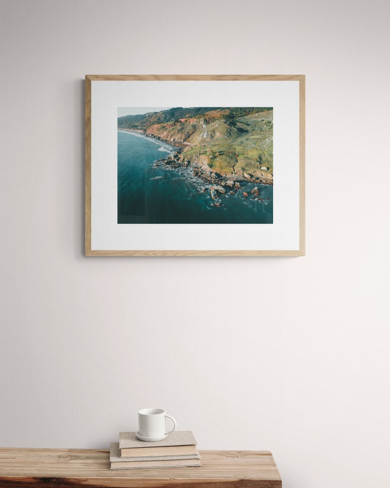 May include: A framed photograph of a rocky coastline with green vegetation and blue water. The photo is in a light wood frame.