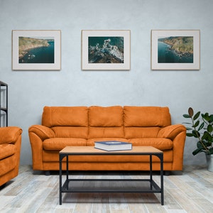 May include: A living room interior with a brown leather sofa, a coffee table, and a shelf unit. Three framed pictures of coastal landscapes hang on the wall above the sofa. A potted plant sits on the floor next to the sofa.