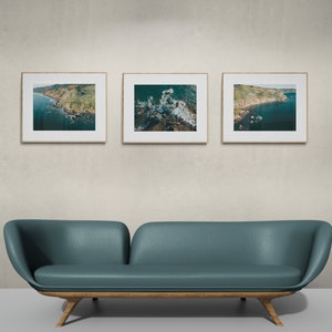 May include: Three framed photographs of aerial views of a coastline with green hills and blue water. The photos are hanging on a light gray wall above a teal blue couch with wooden legs.