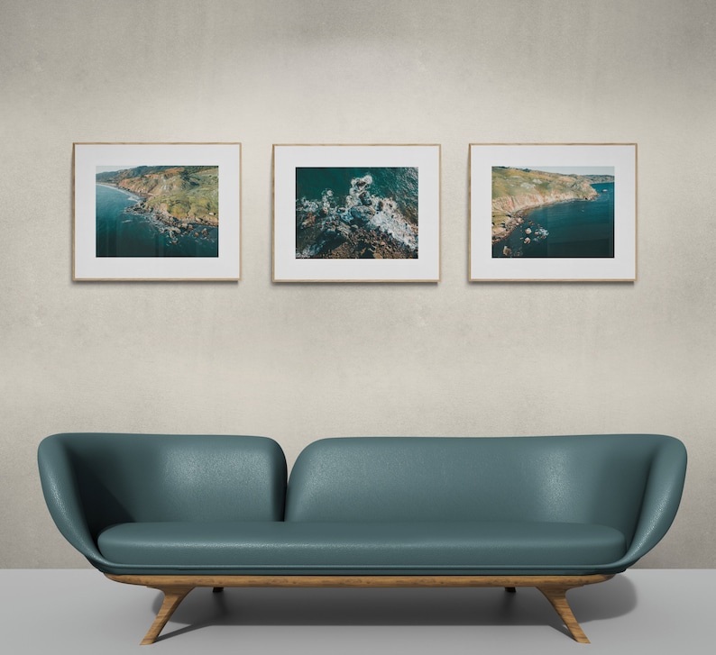 May include: Three framed photographs of aerial views of a coastline with green hills and blue water. The photos are hanging on a light gray wall above a teal blue couch with wooden legs.