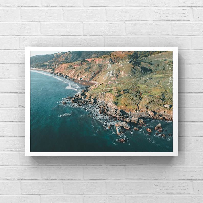 May include: An aerial view of a rugged coastline with green hills, rocky cliffs, and a blue ocean. The water is clear and turquoise, and the waves are crashing against the shore.
