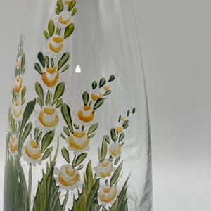 Könnte beinhalten: Eine transparente Glaskaraffe mit handgemalten Blumenmustern. Die Blumen sind weiß und gelb mit grünen Blättern und Stielen. Die Karaffe hat eine schlanke, elegante Form.