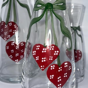 Könnte beinhalten: Klare Glasvasen mit handgemalten roten Herzen und grünen Schleifen. Die Herzen haben weiße Tupfen. Diese dekorativen Vasen eignen sich für den Valentinstag oder andere besondere Anlässe.