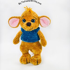 May include: A golden-brown crocheted plush animal with large ears featuring pink inner accents, blue plastic eyes, a dark brown nose, and a blue textured shirt. The text ByTatianaWithLove is positioned above the head of the plush toy.