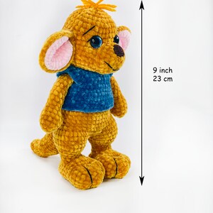 May include: A golden-brown crocheted plush toy with a blue vest, large ears with pink centers, and dark blue eyes. The toy features a small tuft of orange yarn on its head and a dark brown nose. It measures 9 inches in height as indicated by text.