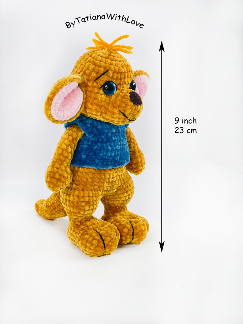 May include: A golden-brown crocheted plush toy with a blue vest, large ears with pink centers, and dark blue eyes. The toy features a small tuft of orange yarn on its head and a dark brown nose. It measures 9 inches in height as indicated by text.