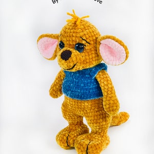May include: A crocheted plush toy of a kangaroo character with golden-brown yarn, large ears with pink inner fabric, dark eyes, a brown nose, and a blue shirt. The text ByTatianaWithLove is visible above the head of the plush toy.