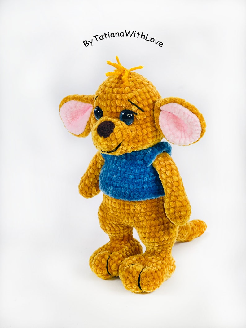 May include: A crocheted plush toy of a kangaroo character with golden-brown yarn, large ears with pink inner fabric, dark eyes, a brown nose, and a blue shirt. The text ByTatianaWithLove is visible above the head of the plush toy.