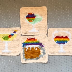May include: Four crocheted drink coasters. Each coaster features a different drink design: a cocktail, a martini, a beer mug, and a wine glass. The drink designs are in rainbow colors. The coasters are square and beige.