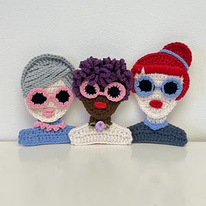 Può includere: Tre figure di donne all'uncinetto con diversi colori e stili di capelli, che indossano occhiali da sole e abiti colorati. Le figure sono realizzate con una varietà di colori e trame di filato.
