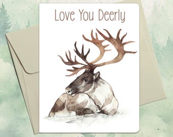 Puede incluir: Una tarjeta de felicitación blanca con una ilustración en acuarela de un reno marrón y blanco descansando en la nieve. El texto "Love You Deerly" está impreso en marrón en la parte superior. Un sobre a juego de color claro está detrás.