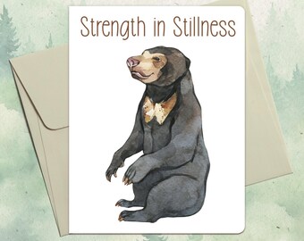 May include: A white greeting card featuring a watercolor illustration of a black sun bear with a tan chest patch. The text Strength in Stillness is printed in brown above the bear. The card is placed on top of a light tan envelope.