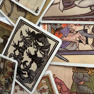 May include: A collection of vintage tarot cards with detailed illustrations. The cards feature images in shades of brown, black, red, and purple. The artwork depicts figures, scenes, and symbols, with white borders. The cards are arranged in a scattered layout.