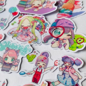 May include: A collection of colourful stickers featuring cartoon characters with different expressions and poses. The stickers are on a white background and include a girl reading a book, a girl with a magnifying glass, a girl with a lollipop, and a girl with a cat.