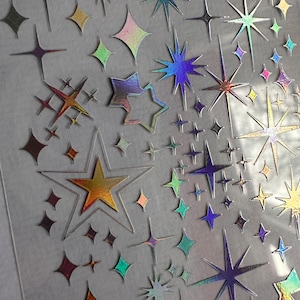 May include: A sheet of iridescent star stickers in various sizes and shapes. The stickers have a holographic effect, displaying colors like gold, blue, green, and purple. The stars include classic five-pointed shapes, diamond shapes, and starburst designs.