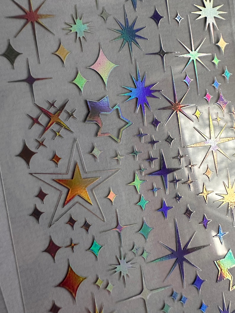 May include: A sheet of iridescent star stickers in various sizes and shapes. The stickers have a holographic effect, displaying colors like gold, blue, green, and purple. The stars include classic five-pointed shapes, diamond shapes, and starburst designs.