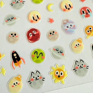 Puede incluir: Hoja de pegatinas con ilustraciones temáticas del espacio. Los diseños incluyen planetas sonrientes, gatos, cohetes, estrellas, lunas y soles. Las pegatinas son de varios colores, incluyendo amarillo, verde, gris, rojo y naranja.