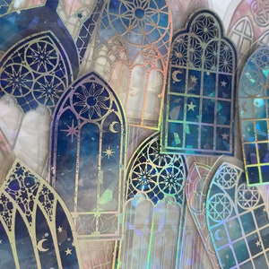 Könnte beinhalten: Eine Sammlung von irisierenden holografischen Aufklebern mit Buntglasfenster-Designs mit Sternen, Monden und geometrischen Mustern. Die Aufkleber sind blau, gold und weiß.