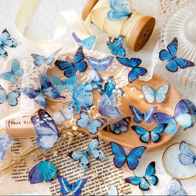 May include: A sheet of clear blue butterfly stickers. The butterflies are various sizes and have a translucent, almost watercolour-like appearance. The stickers are arranged on a white lace background with a wooden spool of ribbon and a piece of paper with text.