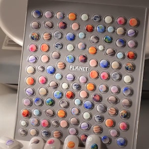 Può includere: Un foglio di adesivi per nail art con disegni di pianeti colorati. Gli adesivi includono vari pianeti con dettagli come anelli e motivi a spirale, in tonalità di blu, arancione, rosa e grigio. La parola "PLANET" è stampata.