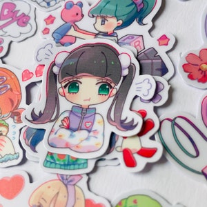 May include: A collection of colourful cartoon stickers featuring characters with different expressions and poses. The stickers are printed on white paper and have a glossy finish.