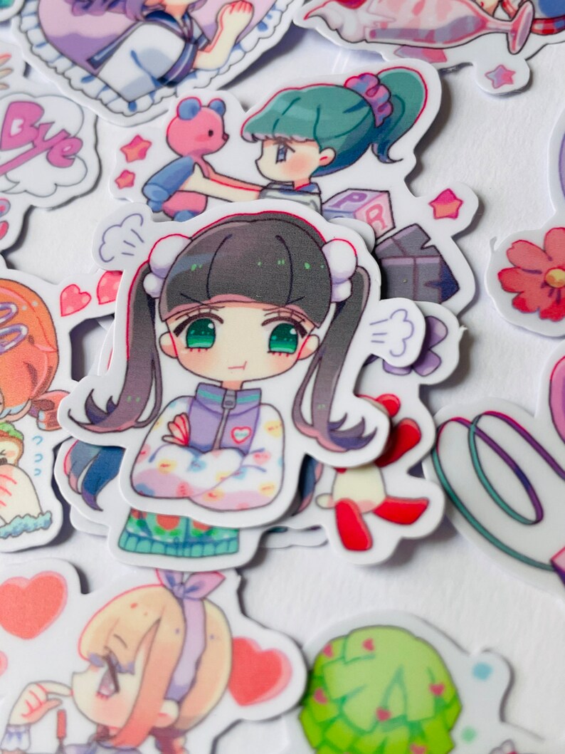 May include: A collection of colourful cartoon stickers featuring characters with different expressions and poses. The stickers are printed on white paper and have a glossy finish.