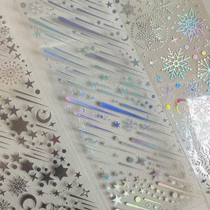 May include: Clear, iridescent sticker sheets featuring various celestial and winter-themed designs. The stickers include stars, snowflakes, shooting stars, and crescent moons in silver and holographic finishes. Ideal for scrapbooking, card making, or other craft projects.