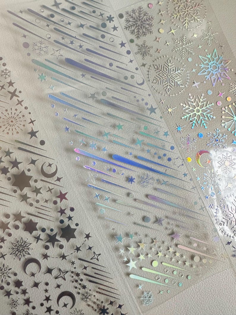 May include: Clear, iridescent sticker sheets featuring various celestial and winter-themed designs. The stickers include stars, snowflakes, shooting stars, and crescent moons in silver and holographic finishes. Ideal for scrapbooking, card making, or other craft projects.