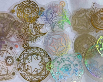 May include: A collection of iridescent holographic stickers featuring various circular designs with intricate patterns, including stars, moons, and geometric shapes. The stickers are transparent and have a gold foil finish.