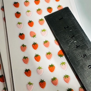 May include: A sheet of clear sticker paper with multiple red and pink strawberry stickers. The stickers are approximately 1/2 inch in diameter. A ruler is in the background.