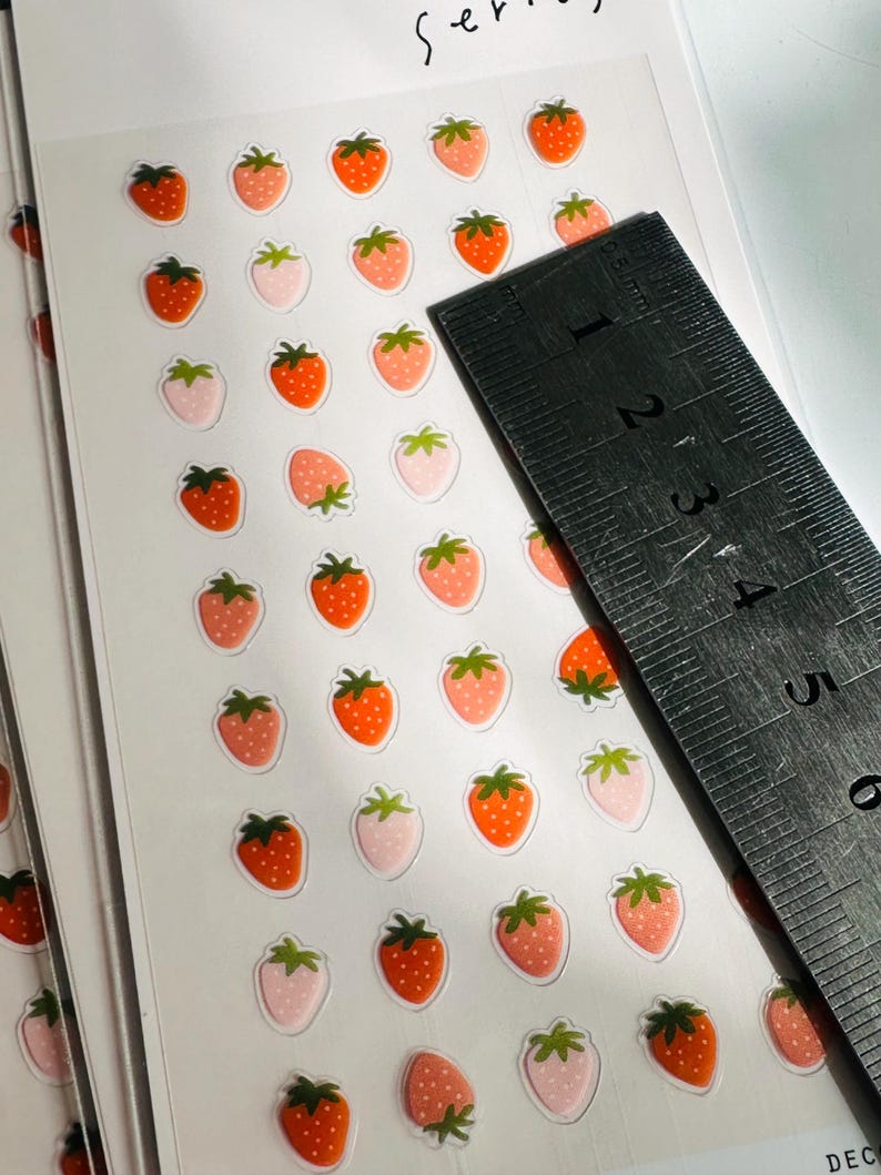 May include: A sheet of clear sticker paper with multiple red and pink strawberry stickers. The stickers are approximately 1/2 inch in diameter. A ruler is in the background.