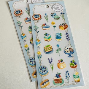 May include: Two sticker sheets with watercolour illustrations of cakes, tarts, and pastries, with floral accents. The stickers are arranged on a light blue background with the text "C'est bon sticker".