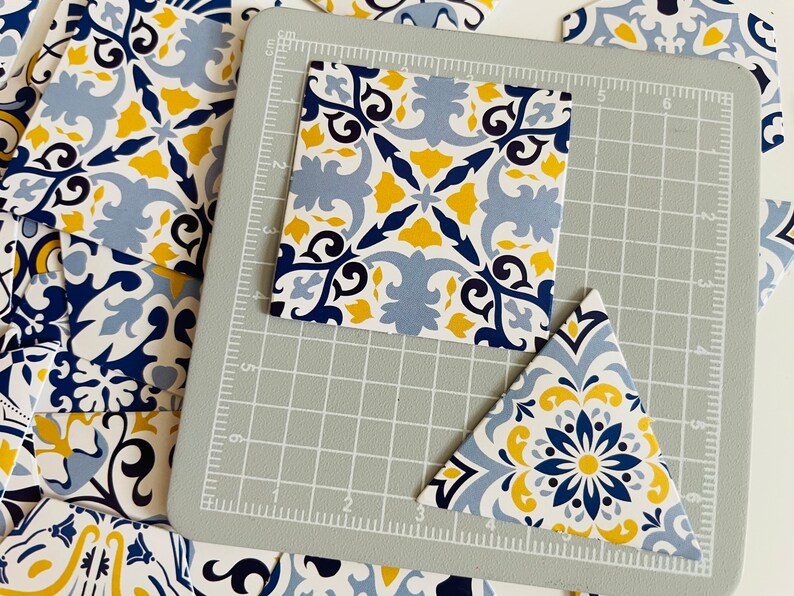 May include: A close-up of two blue and yellow patterned tiles, one square and one triangular, on a gray cutting mat. The tiles have a floral design with blue, yellow, and white accents.