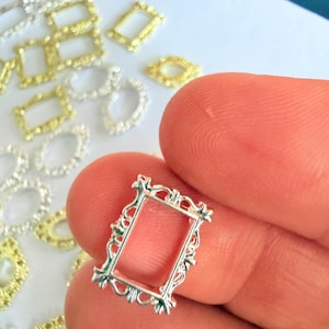 Könnte beinhalten: Ein kleiner, rechteckiger silberfarbener Metallrahmen-Anhänger mit verzierten, schnörkelartigen Details an den Rändern. Der Rahmen ist in der Mitte hohl und weist ein dekoratives, filigranes Randdesign auf. Mehrere ähnliche Rahmen-Anhänger in Silber- und Goldtönen sind im Hintergrund verstreut.