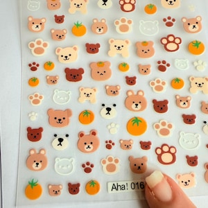 以下が含まれることがあります： 茶色、ベージュ、白のクマのデザイン、オレンジ色の果物、足跡が描かれたステッカーシート。ステッカーは光沢仕上げで、「Aha! 016」の文字が含まれています。
