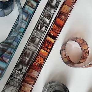 May include: Three rolls of washi tape with a film strip design. The tapes feature different colour schemes and images, including a blue and white tape with cityscapes, a black and white tape with a more abstract design, and a brown and orange tape with sunset scenes.