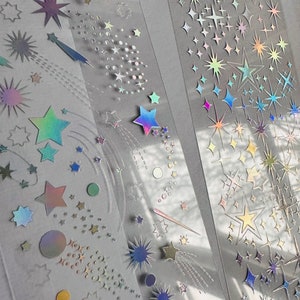 May include: Clear tape rolls featuring holographic star and comet designs. The stars vary in size and color, with iridescent and metallic finishes. Some stars have trailing lines, creating a shooting star effect. Ideal for scrapbooking and crafts.