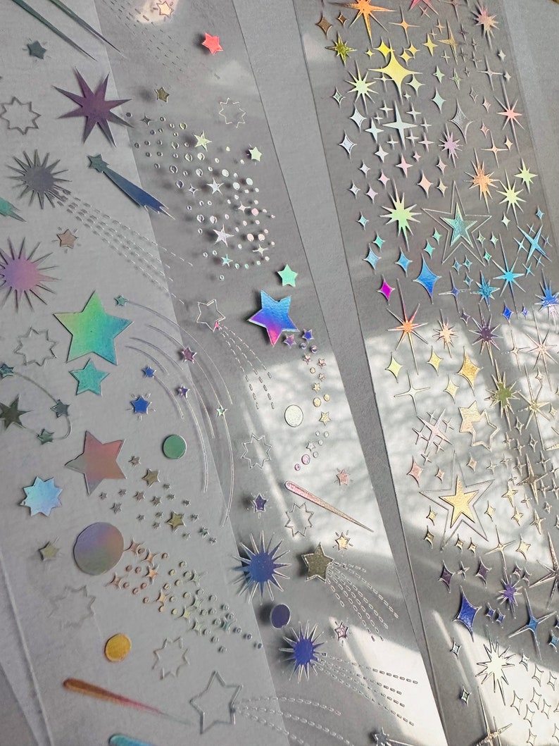 May include: Clear tape rolls featuring holographic star and comet designs. The stars vary in size and color, with iridescent and metallic finishes. Some stars have trailing lines, creating a shooting star effect. Ideal for scrapbooking and crafts.