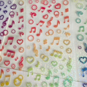 Op de afbeelding: Vellen kleurrijke, glinsterende stickers met muzieknoten, G-sleutels, harten en trouwringen. De stickers zijn in een regenboog van kleuren, waaronder paars, rood, oranje, geel, groen en blauw. Perfect voor scrapbooking en knutselen.