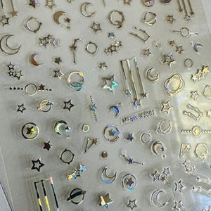 May include: A sheet of holographic stickers with celestial designs. The stickers include stars, moons, planets, and constellations in silver and iridescent finishes. The sheet has the text "MYSW-A1002".