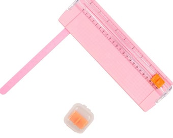 Puede incluir: Un cortador de papel rosa con una cuchilla de plástico transparente y una regla marcada en centímetros. Viene con un pequeño reemplazo de cuchilla naranja en una caja de plástico transparente.