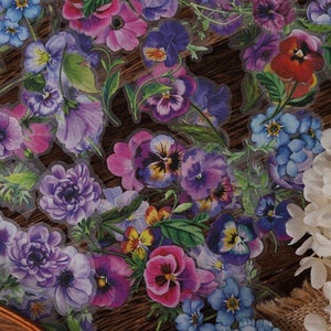 May include: A sheet of clear stickers featuring various watercolor floral designs. The stickers feature purple, blue, and pink flowers with green leaves. The flowers are arranged in a random pattern on the sheet.