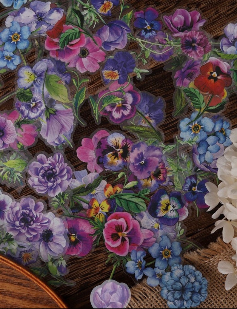 May include: A sheet of clear stickers featuring various watercolor floral designs. The stickers feature purple, blue, and pink flowers with green leaves. The flowers are arranged in a random pattern on the sheet.