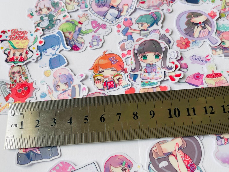 May include: A sheet of colourful stickers featuring cartoon characters with different expressions and poses. The stickers are arranged on a white surface with a ruler measuring 13 centimetres.