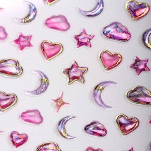 May include: A collection of pink and purple stickers featuring celestial shapes. The stickers include hearts with gold trim, crescent moons, stars, and cloud shapes. The stickers have a glittery, translucent appearance.