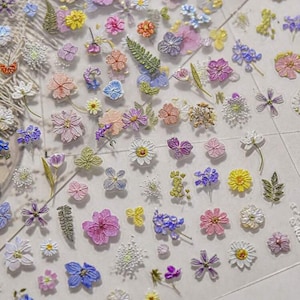 Peut inclure: Une collection de petites décorations florales colorées de différentes formes. L'assortiment comprend des marguerites, des pensées et d'autres fleurs dans les tons de rose, de violet, de jaune et de blanc. Certains ont des paillettes et des feuilles vertes.