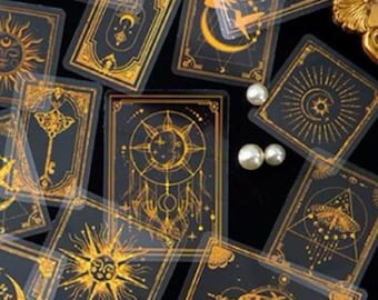 May include: A collection of black and gold tarot cards featuring celestial and mystical designs. The cards display suns, moons, keys, and dreamcatcher illustrations. Scattered pearls add a decorative touch to the arrangement.